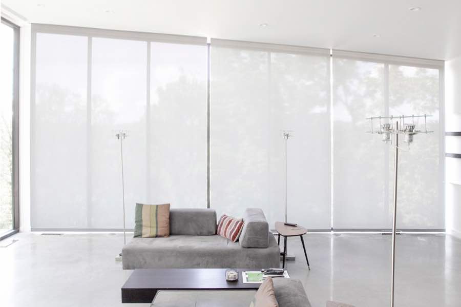 Motorized window shades make controlling natural light effortless in your Riverside great room. White motorized shades on a wall of tall great room windows