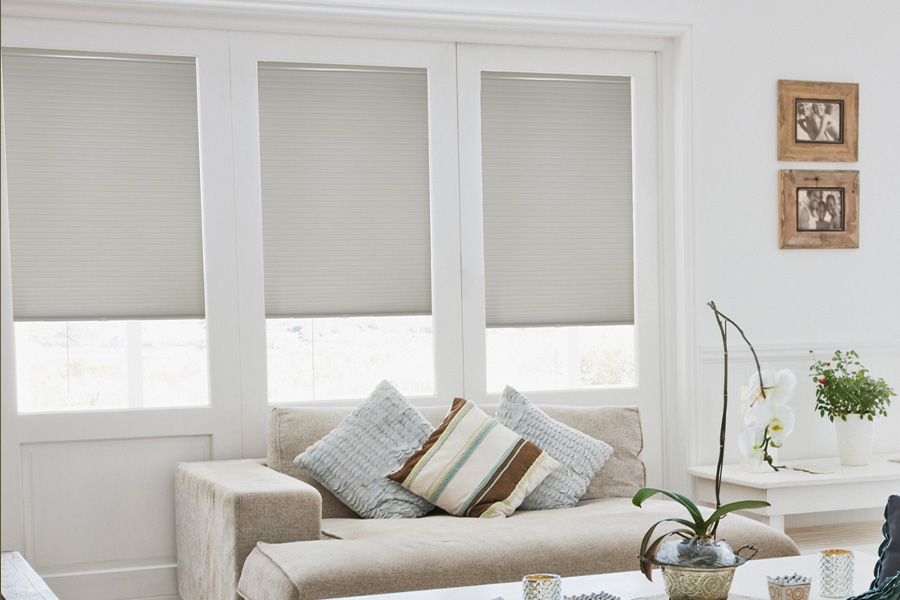 Cellular shades are energy-conservative for your living spaces.  Light gray cellular shades on three separate windows in large light wood and beige colored living room.